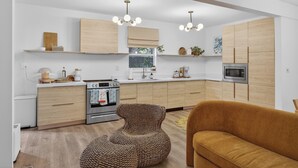 A modern kitchen with wooden cabinets, a built-in oven, and a microwave. There is a brown woven chair and a mustard-colored sofa in the living area. - Little Havana Cabana – Retro Surf-Lodge Hideaway (Miami)