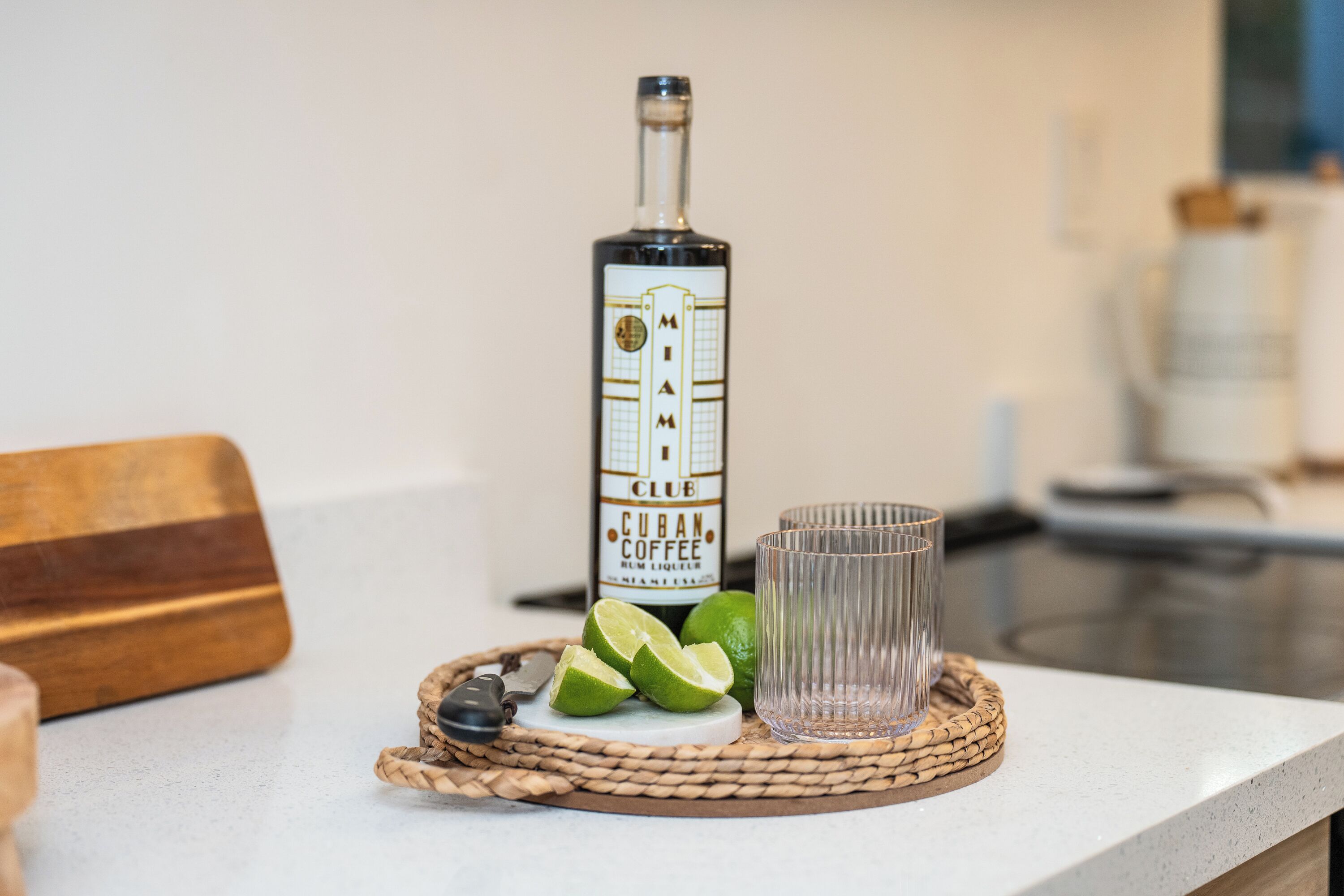 A bottle of Cuban Coffee rum liqueur, lime wedges, and a glass on a woven coaster on a kitchen counter.