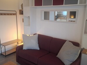 A living room with a red sofa, two white cushions, a wooden side table, and a built-in shelf unit. - New studio in Corse-du-Sud (Figari)