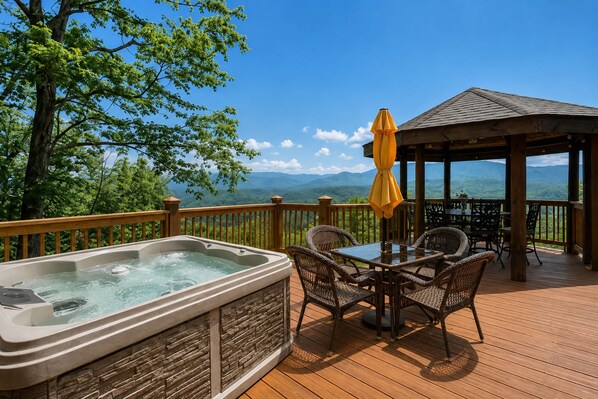Un bain à remous sur une terrasse en bois, agrémentée de fauteuils en osier et d’une table, offrant une vue sur un paysage montagneux.
