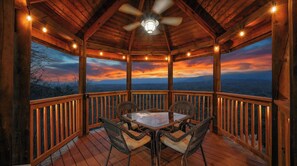 Une terrasse en bois, agrémentée d’une table et de chaises, entourée de rampes en bois, et offrant une vue panoramique sur un paysage magnifique au coucher du soleil.