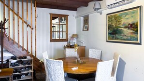 Un comedor con una mesa de madera, sillas blancas con diseño, un cuadro en la pared y una escalera con barandillas de madera.