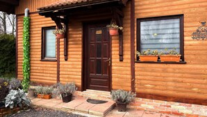 Una cabaña de madera con puerta marrón oscuro, dos ventanas y un pequeño porche con macetas.