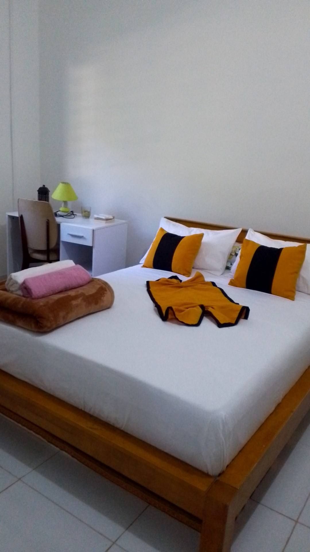 A neatly made bed with pillows and a blanket, a desk with a lamp, and a chair in a room with white walls.