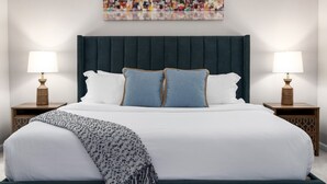 Een net opgemaakt bed met witte lakens, twee kussens en een plaid. Er zijn twee nachtkastjes met lampen en een groot abstract schilderij erboven.