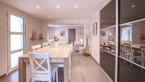 A modern dining area with a wooden table and white chairs, a sofa, and a large window. - L'Oustal Occitan: Idyllic Family Escape with Private Pool (Saint-Privat-des-Vieux)