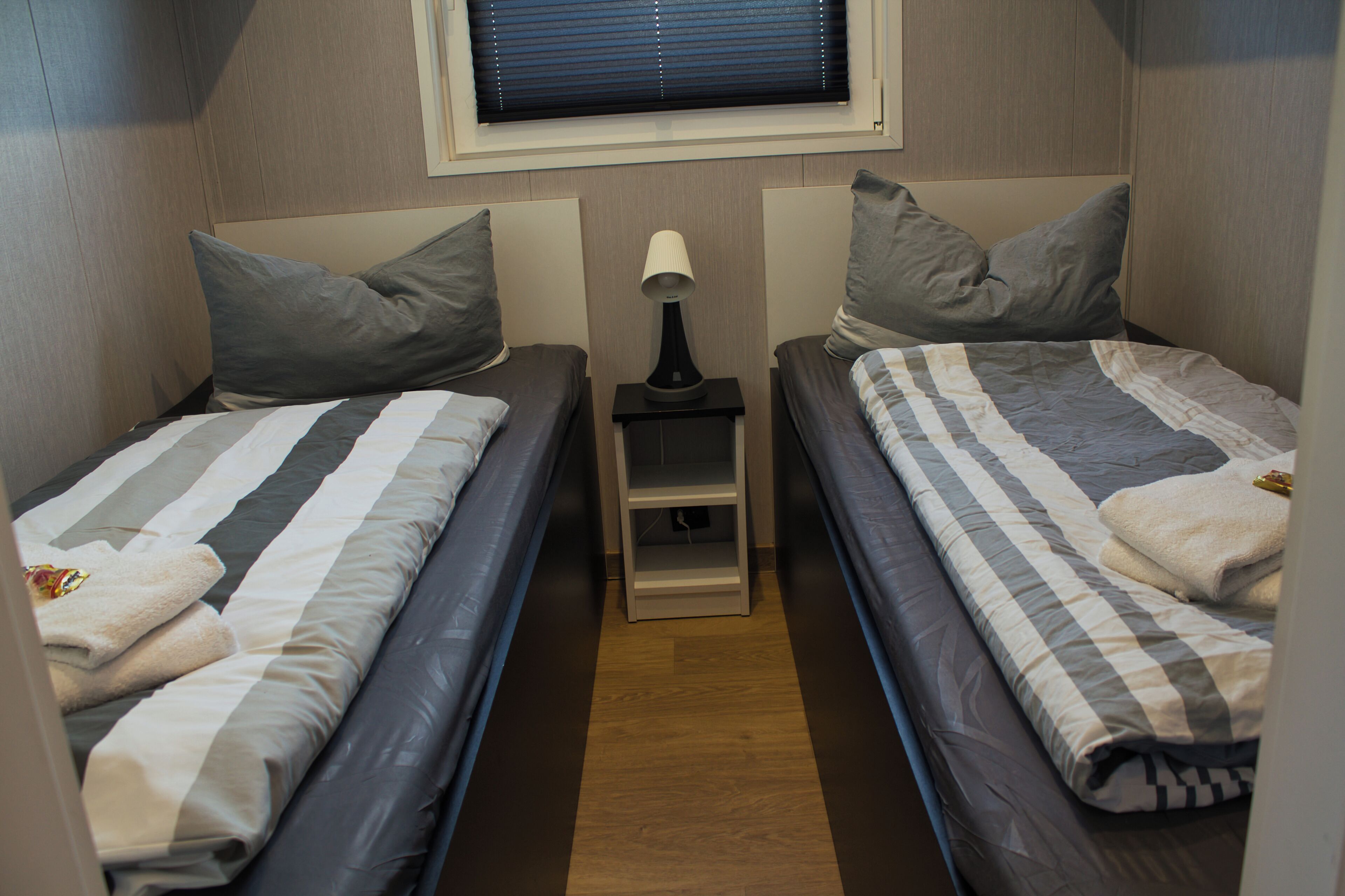 Une petite chambre compacte, équipée de deux lits simples, d’une table de chevet avec une lampe et d’une fenêtre avec des stores.