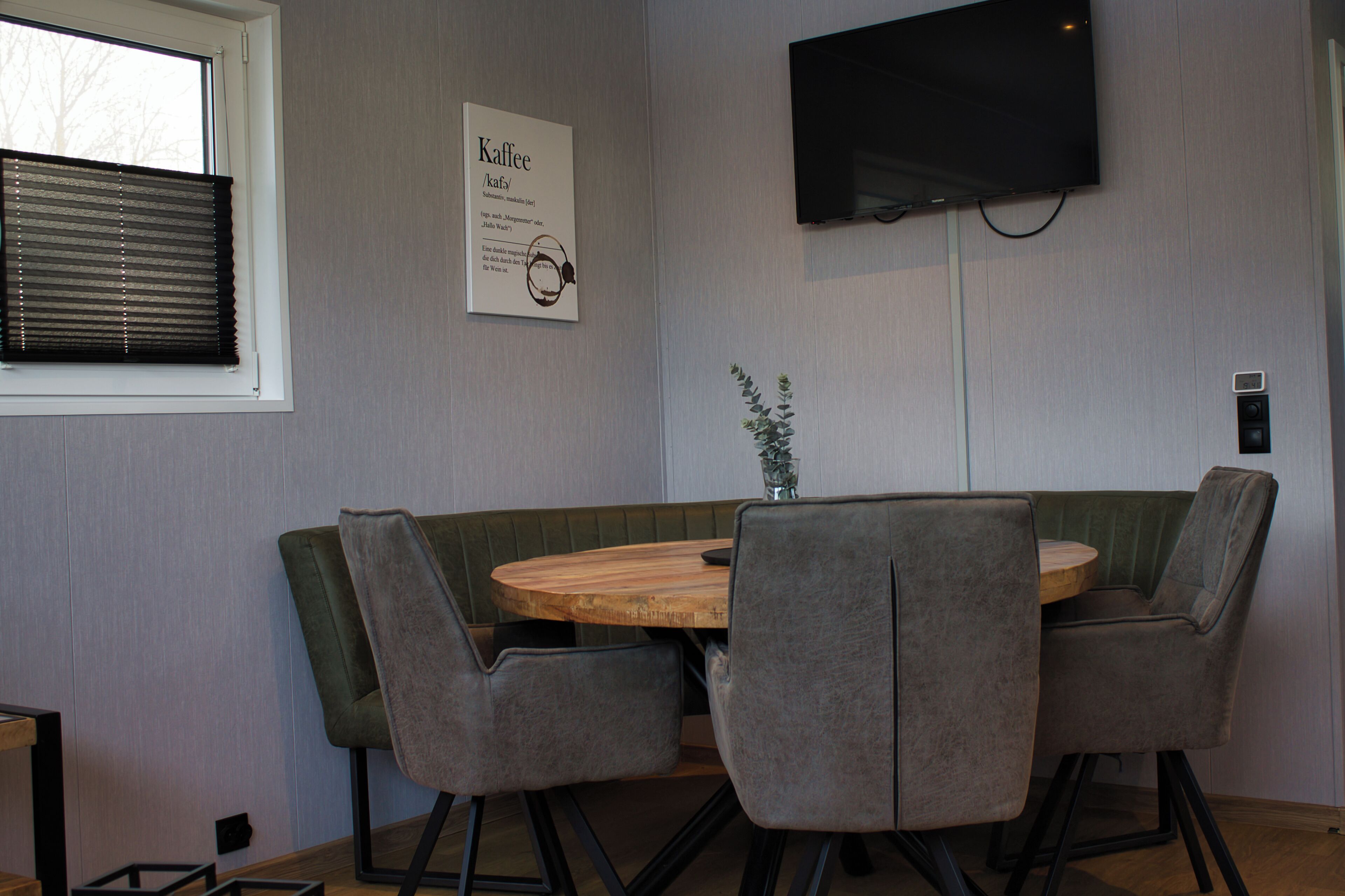 Un coin repas comprenant une table ronde en bois, quatre chaises grises à revêtement rembourré, un téléviseur fixé au mur et une fenêtre avec des stores.