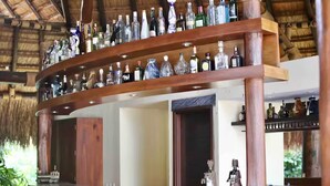 Uma área de bar com várias prateleiras de garrafas de bebidas, balcão de madeira e teto de palha.