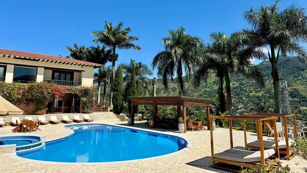 Uma área de piscina com pérgola de madeira e espreguiçadeiras, cercada por palmeiras e um cenário de montanhas.