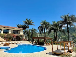 Zona de piscina con una pérgola de madera y sillas de descanso, rodeada de palmeras y un telón de fondo de montañas.