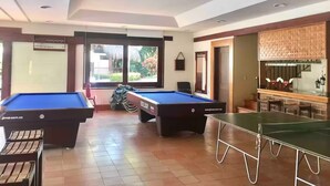 Uma área de piscina com duas mesas de bilhar, uma mesa de tênis de mesa e um bar com fundo de madeira.