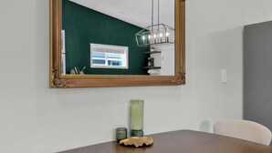 Et spiseområde med et træbord, en grøn vase og en dekorativ skak. Et stort spejl hænger over bordet og afspejler et vindue samt en loftlampe.