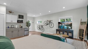 Et moderne soveværelse med en stor seng, et lille bord og et fjernsyn. Der er et tekøkken med skabe og en mikrobølgeovn. To cykler er monteret på væggen.