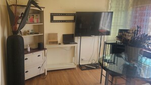 Een woonkamer met een flatscreen televisie, een glazen eettafel, een wit kastje en een zwarte vaas met decoratieve bladeren.