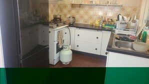 Een kleine keuken met een zwarte koelkast, een oude kookplaat, een gasfles en een spoelbak.