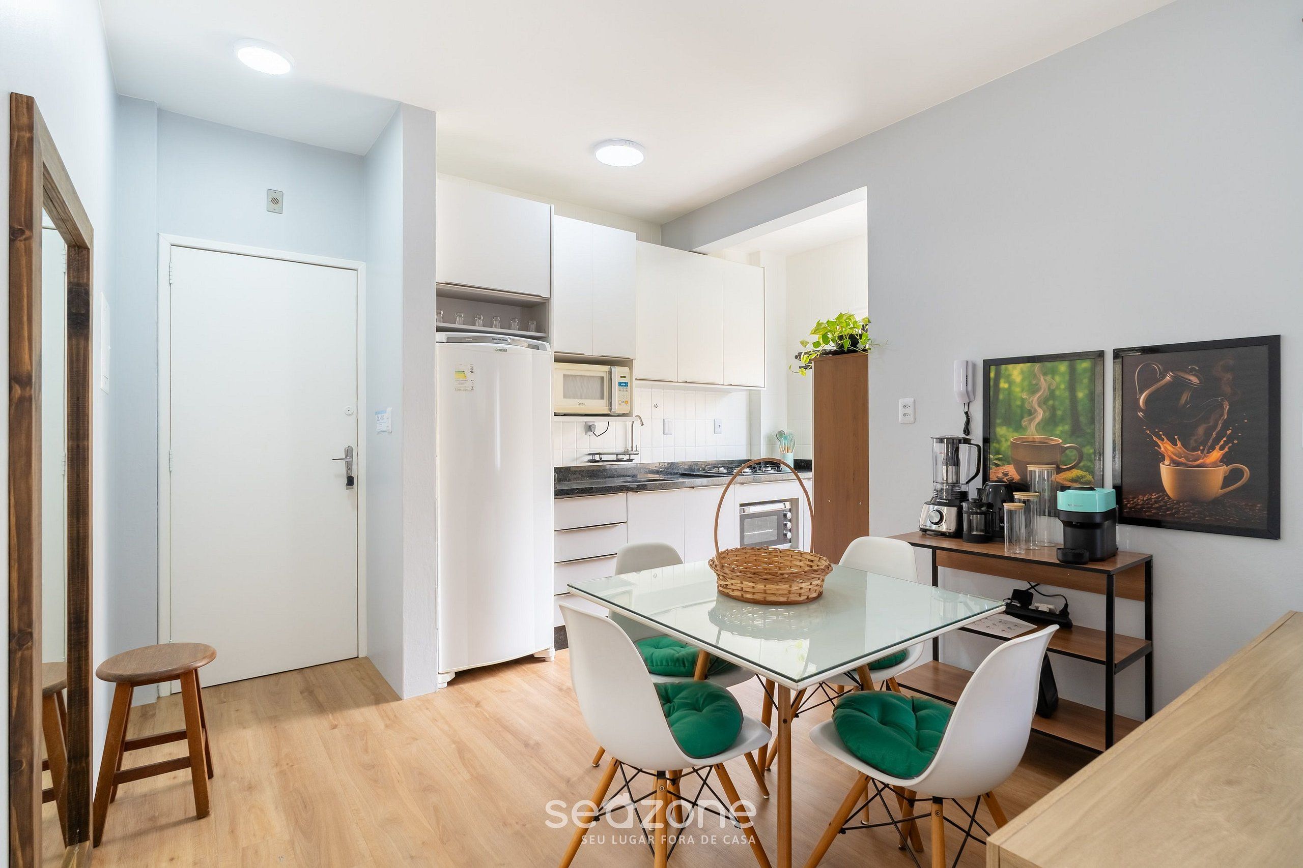 A modern kitchen with white cabinets, a microwave, and a coffee machine. There is a dining area with a glass table and white chairs.