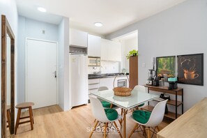 A modern kitchen with white cabinets, a microwave, and a coffee machine. There is a dining area with a glass table and white chairs. - 1-br APT w/ Bbq, 800m From the Sea Dfc0101 (Florianópolis)