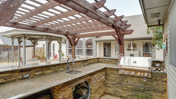 Een buitenkeuken met een stenen aanrechtblad, een ingebouwde grill en een pergola.