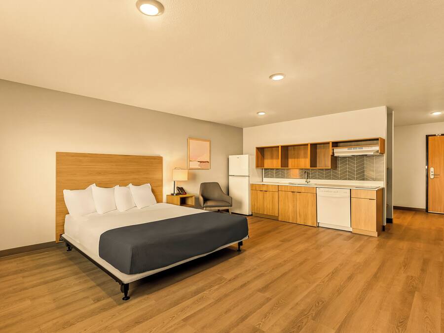 A hotel room with a bed, a desk, a chair, a kitchenette, and a door.