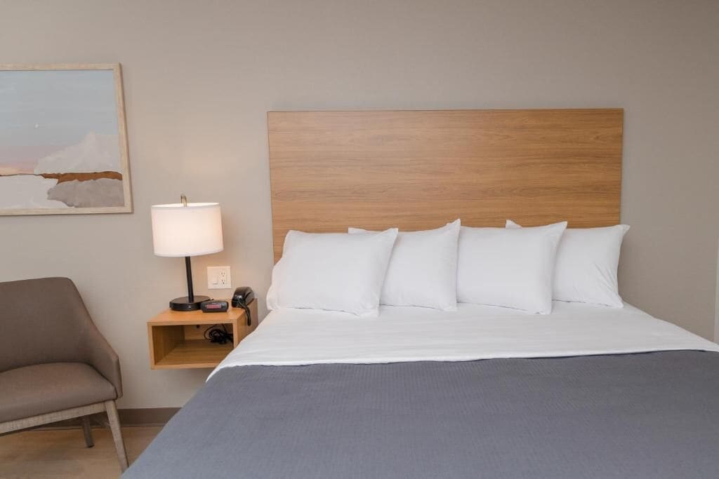 Una habitació d'hotel amb un capçal de llit de fusta, un llit amb roba de llit blanca, una manta grisosa, una làmpada de nit, una cadira i un quadre al mur.
