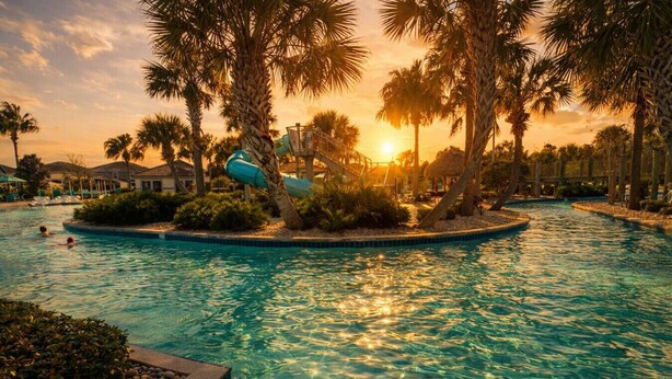 Una piscina con tobogán de agua, rodeada de palmeras y un atardecer.