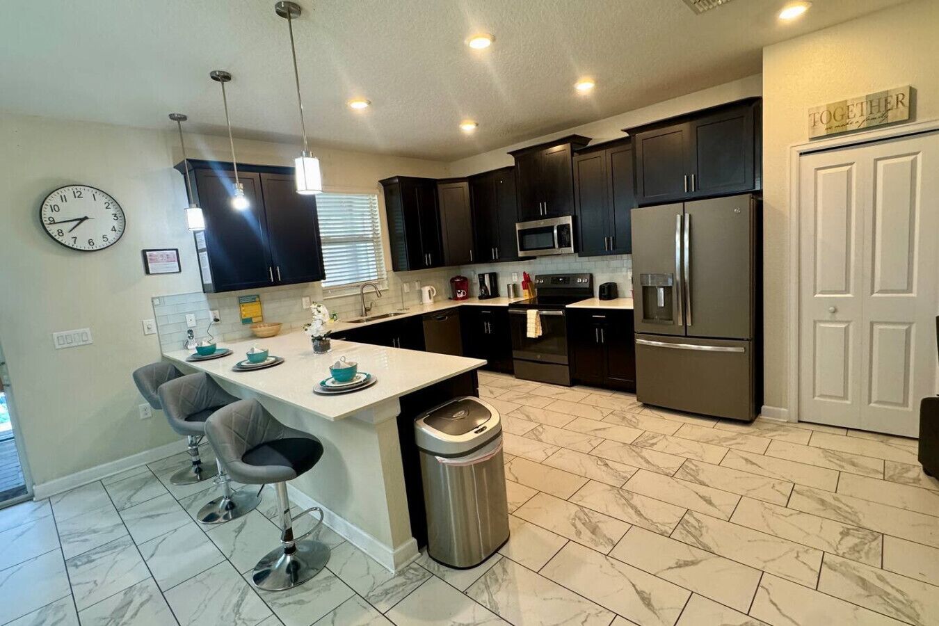 A modern kitchen with dark cabinets, stainless steel appliances, a marble-tiled floor, and a clock on the wall.