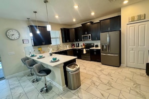 A modern kitchen with dark cabinets, stainless steel appliances, a marble-tiled floor, and a clock on the wall. - The Grand Retreat at ChampionsGate w/pool (Four Corners)
