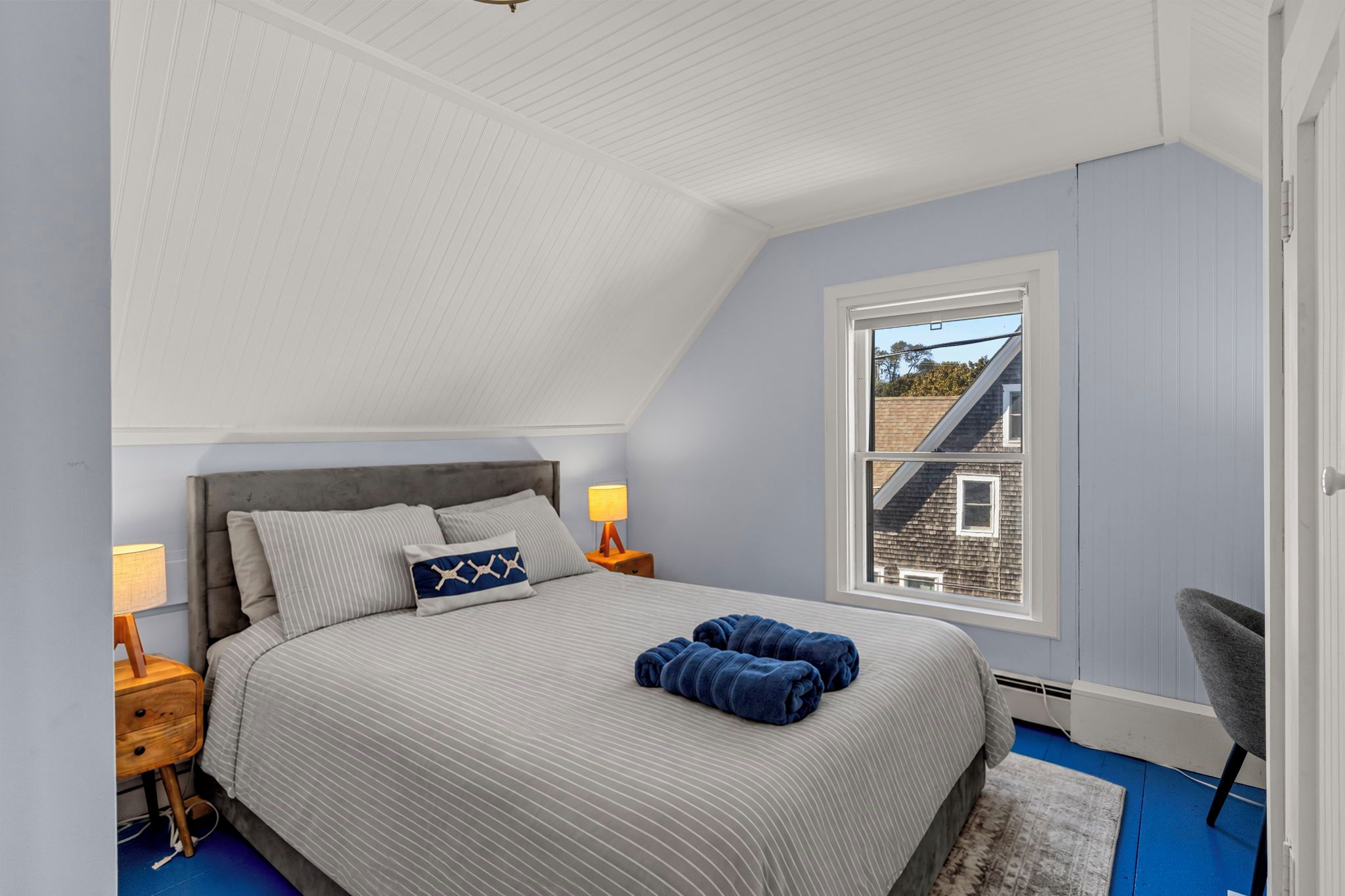Un dormitorio con techo inclinado, una cama con ropa de cama a rayas, dos mesitas de noche con lámparas, una ventana con vista a una casa y una silla.