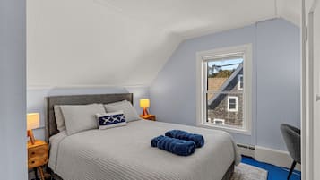 Un dormitorio con techo inclinado, una cama con ropa de cama a rayas, dos mesitas de noche con lámparas, una ventana con vista a una casa y una silla.