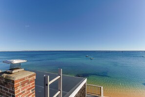 Vista despejada de una playa con aguas tranquilas, un muelle distante y una estructura de ladrillo con una chimenea.