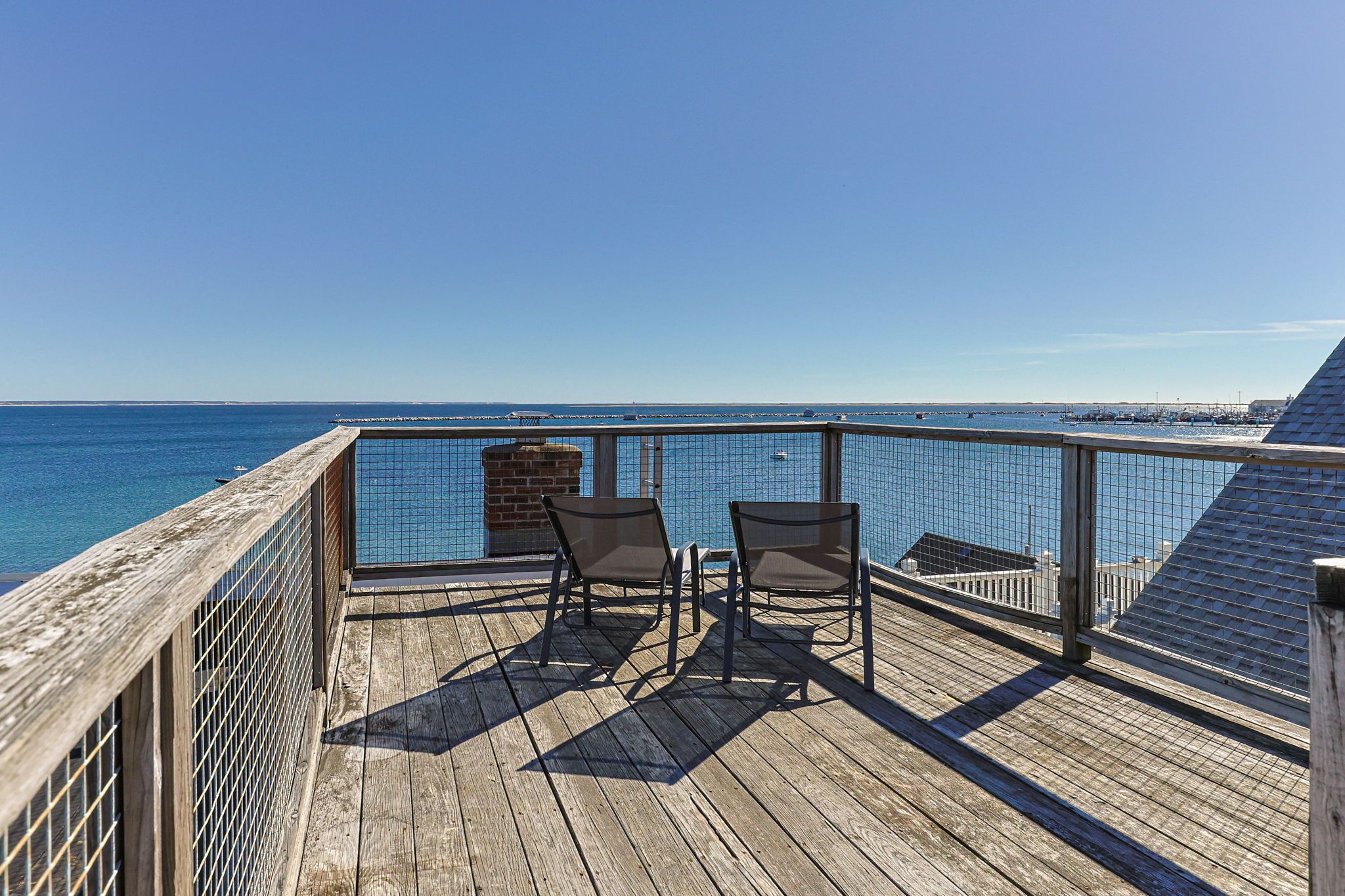 Een houten terras met twee stoelen gericht op een heldere blauwe zee, een bakstenen gebouw en een metalen reling.