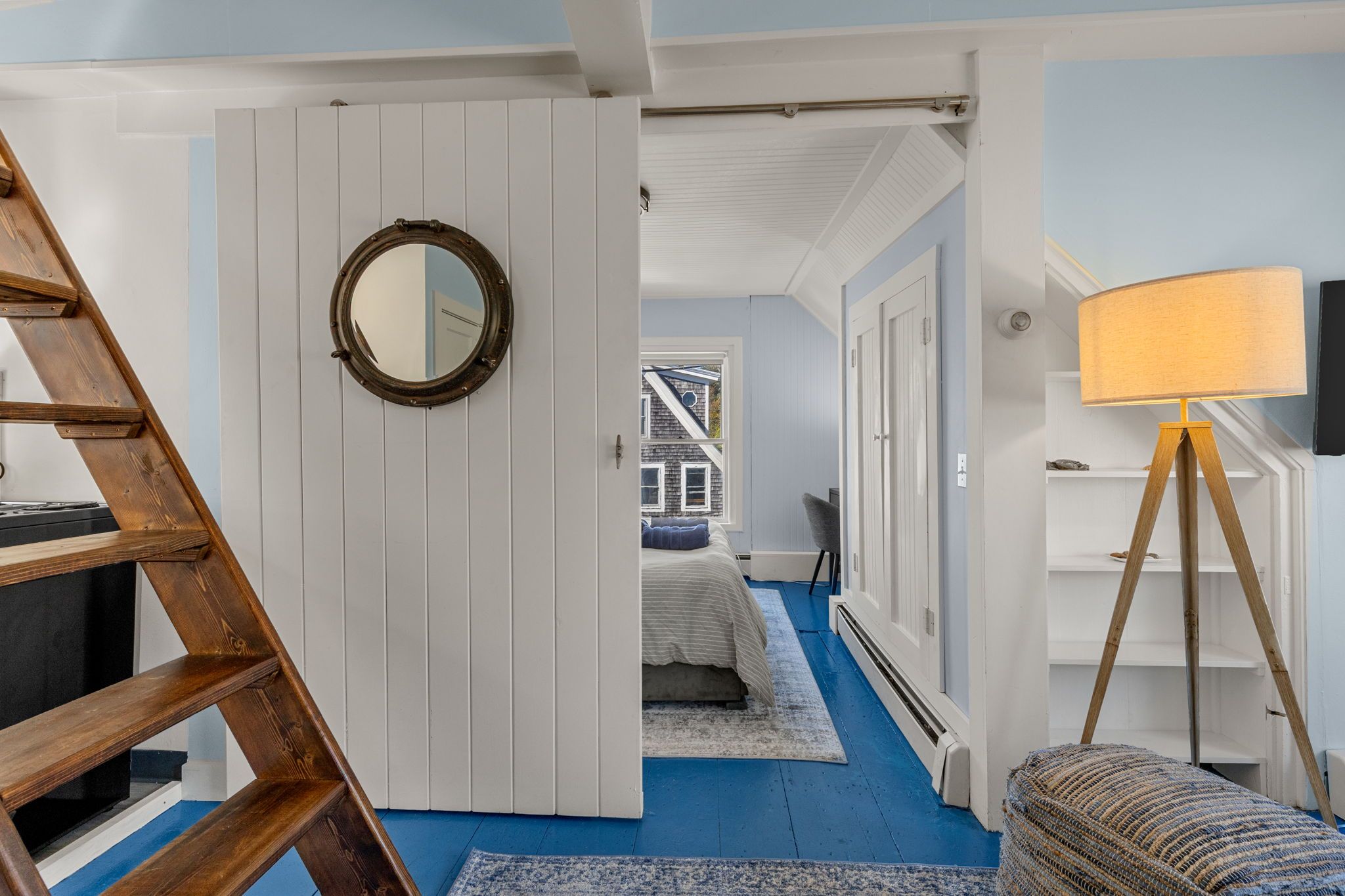 A room with a blue carpet, a wooden ladder, a round mirror, a bed with a blue headboard, a white door, and a lamp.
