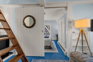 Una habitación con alfombra azul, una escalera de madera, un espejo redondo, una cama con cabecera azul, una puerta blanca y una lámpara.