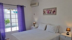 A bedroom with a large bed, two bedside tables, a ceiling light, an air conditioning unit, and a window with purple curtains. - Belsol House (Altura)