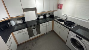 A compact kitchen with white cabinets, a black countertop, a built-in dishwasher, and a window with blinds. - A Home From Home (Crawley)