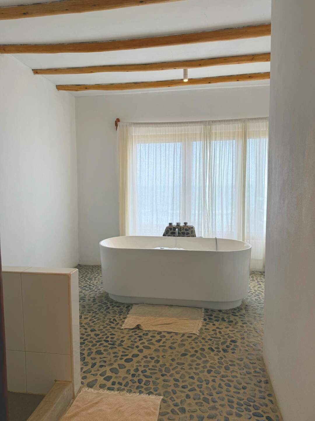 Un baño con una bañera independiente, una ventana con cortinas translúcidas y un suelo con diseño de piedras.