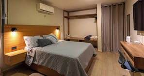 Ein ordentlich eingerichtetes Hotelzimmer mit Bett, Nachttischen, Schreibtisch, Stuhl, Fernseher und Klimaanlage.