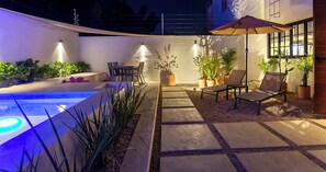 Un patio exterior bien iluminado con piscina, zona de estar y plantas en macetas.