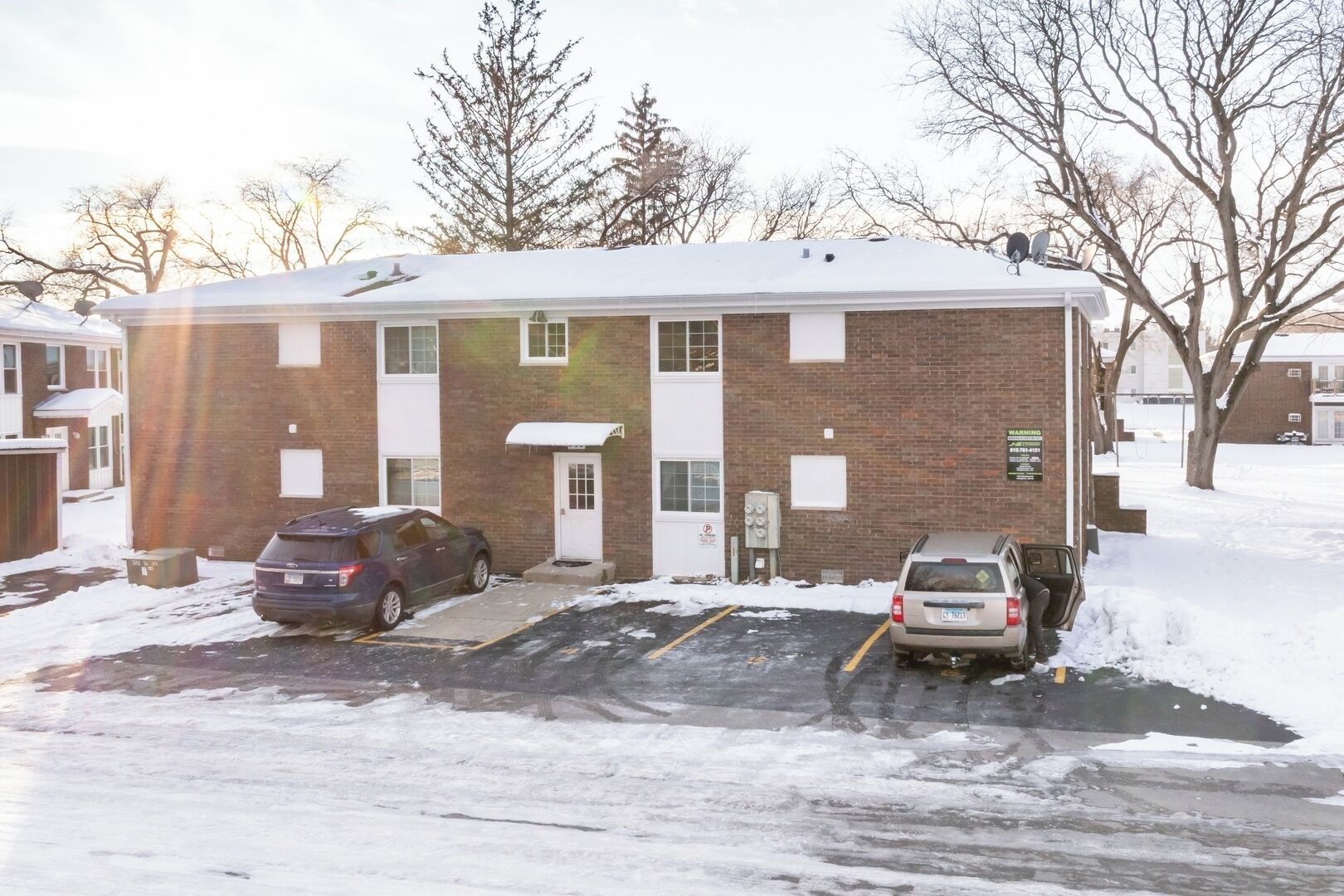A two-story brick building with a snow-covered roof and a parking lot with two cars.