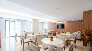 A modern hotel room with multiple round tables and chairs, a flat-screen TV mounted on the wall, and large windows with curtains. - Fully Equipped Apts With Leisure Minutes From the Beach Hfos (Salvador)