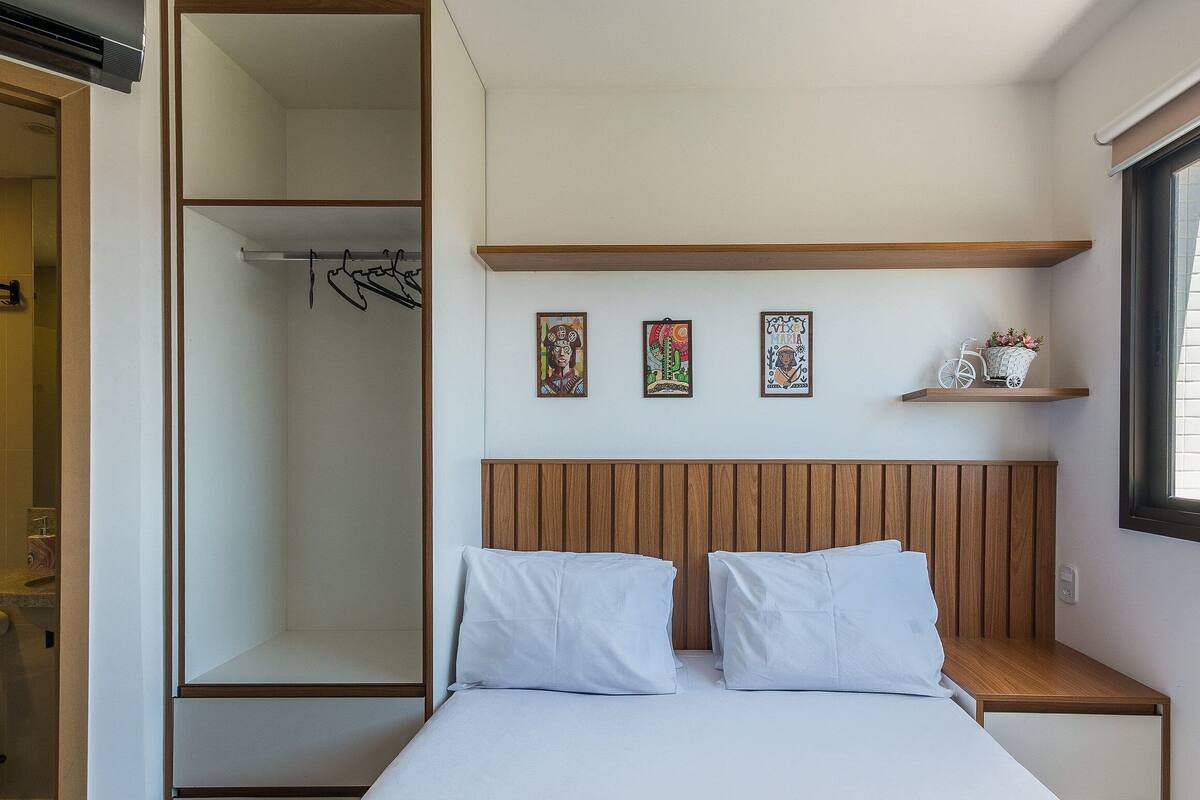 A bedroom with a bed, a wooden headboard, a wardrobe, a window with a city view, and three wall-mounted artworks.