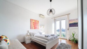 A modern bedroom with a bed, bedside tables, a sofa, and a view of the city through a large window. - Aurelia Suite – Spacious W/ Ac & Danube View (Budapest)