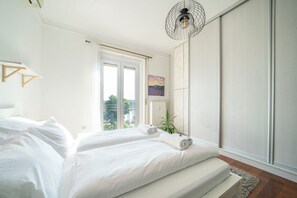 Een slaapkamer met een groot bed, een raam met gordijnen, een kamerplant en een wandlamp.