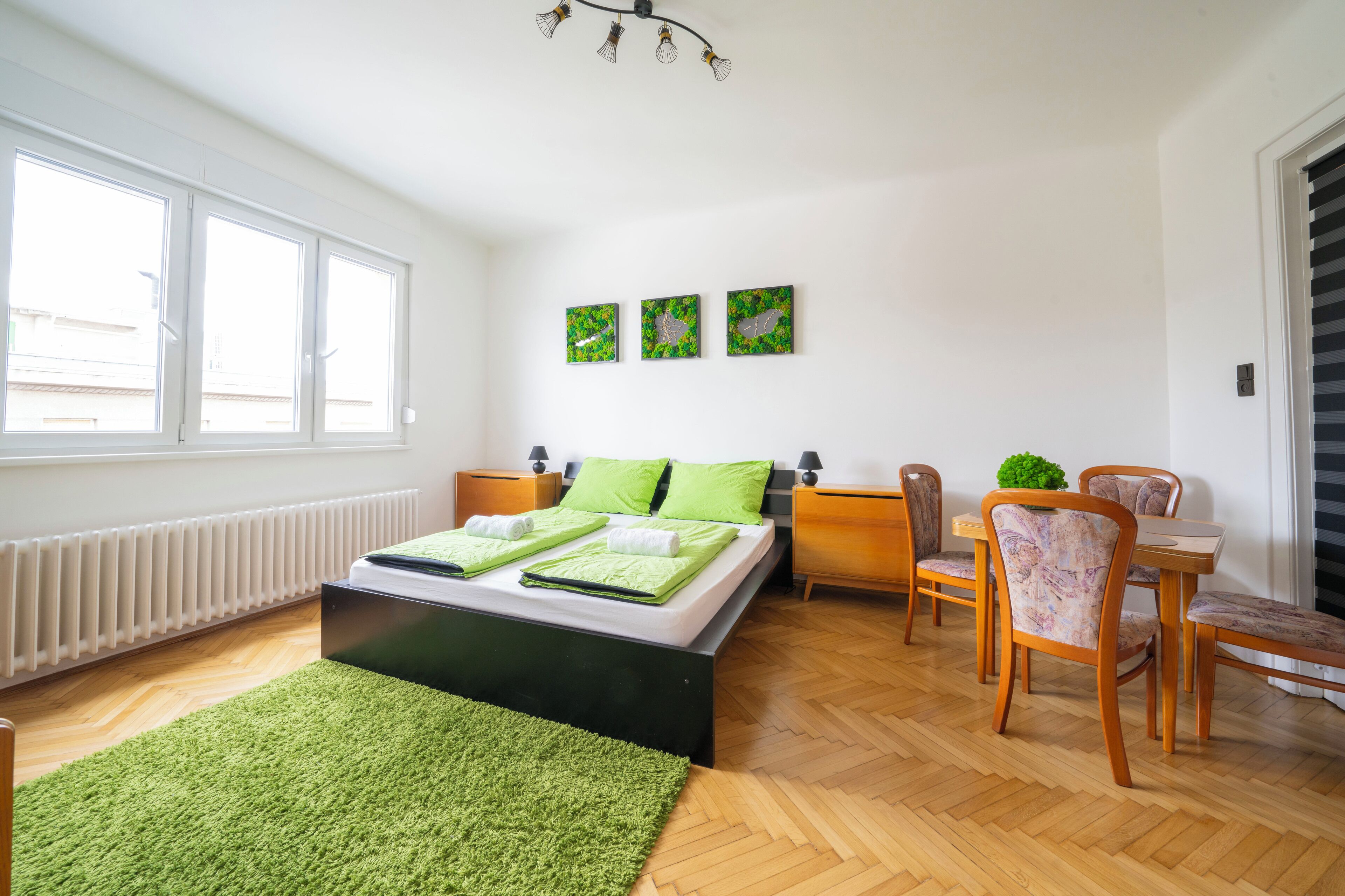 A bedroom with a bed, a desk, and a chair. There are three windows and a radiator.