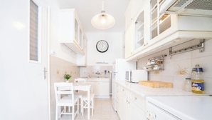 Een lichte keuken met witte kastjes, een eettafel met stoelen, een klok aan de muur en een magnetron op het aanrecht.