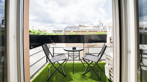 Een balkon met twee stoelen en een kleine tafel, omringd door kunstgras, met uitzicht op een stadsbeeld met gebouwen en een bewolkte lucht.