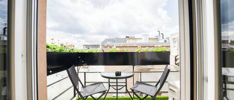 A balcony with two chairs and a small table, surrounded by artificial grass, overlooking a cityscape with buildings and a cloudy sky.