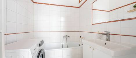 A modern bathroom with white tiles, a built-in washing machine, a bathtub, and a vanity with a mirror.