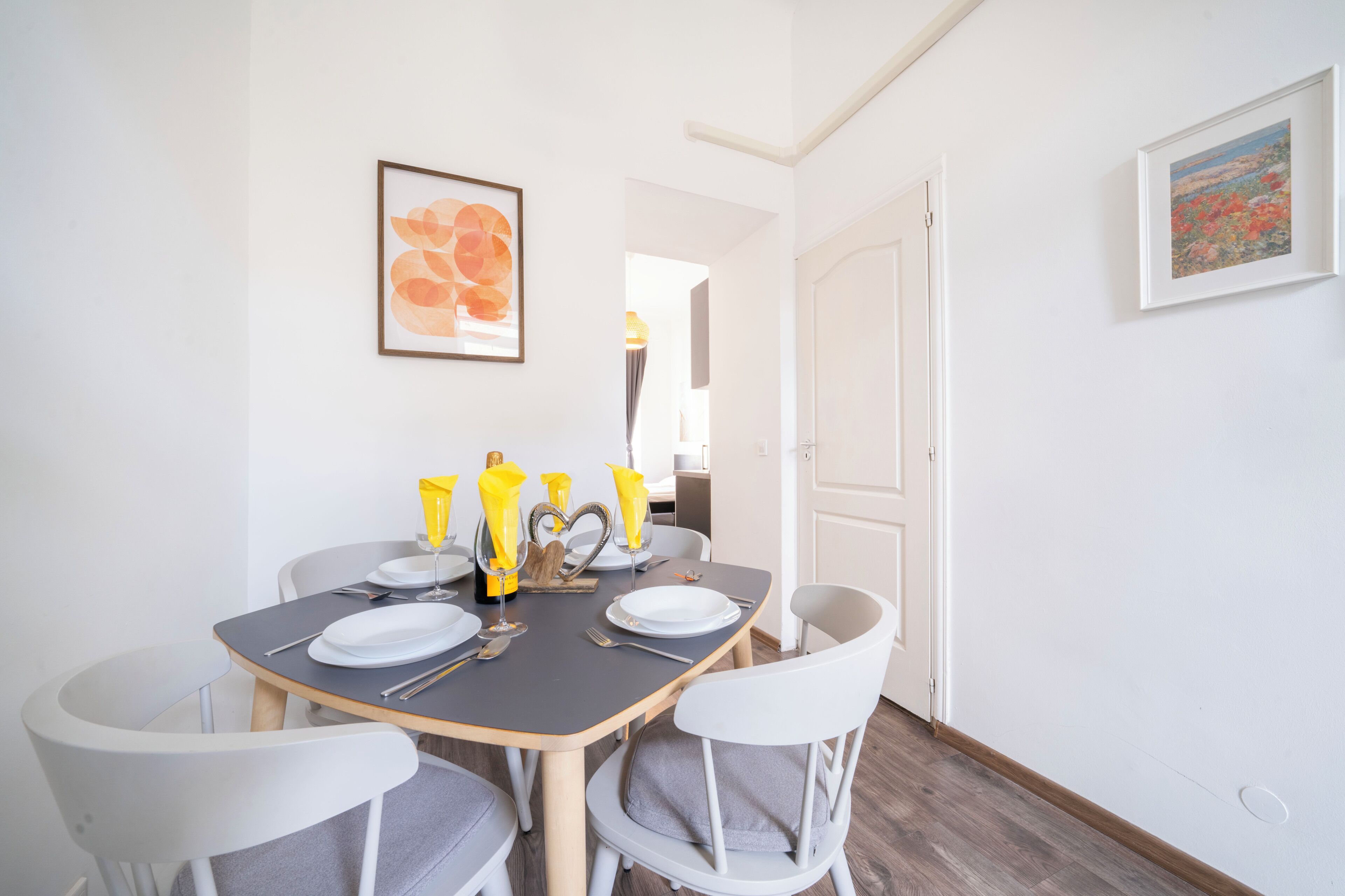 A dining area with a table set for four, chairs, a framed picture on the wall, and a door leading to another room.